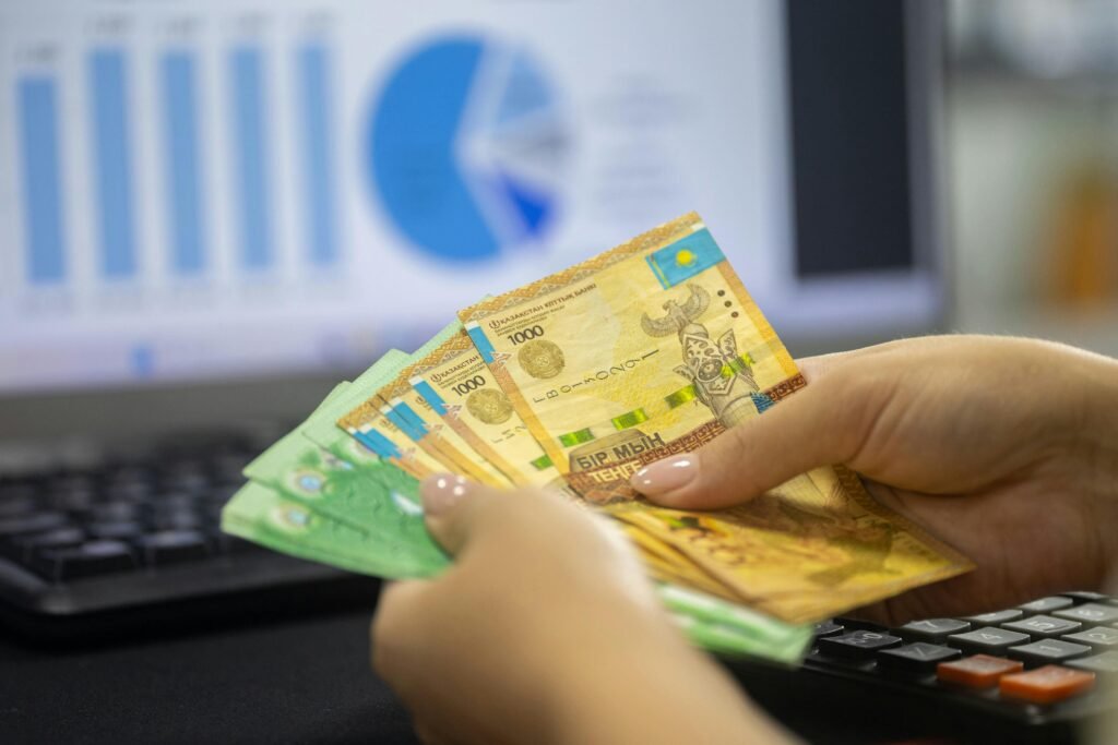 pexels-photo-33175661-33175661 Hands holding Kazakhstan tenge bills with financial chart and graphs on computer screen.