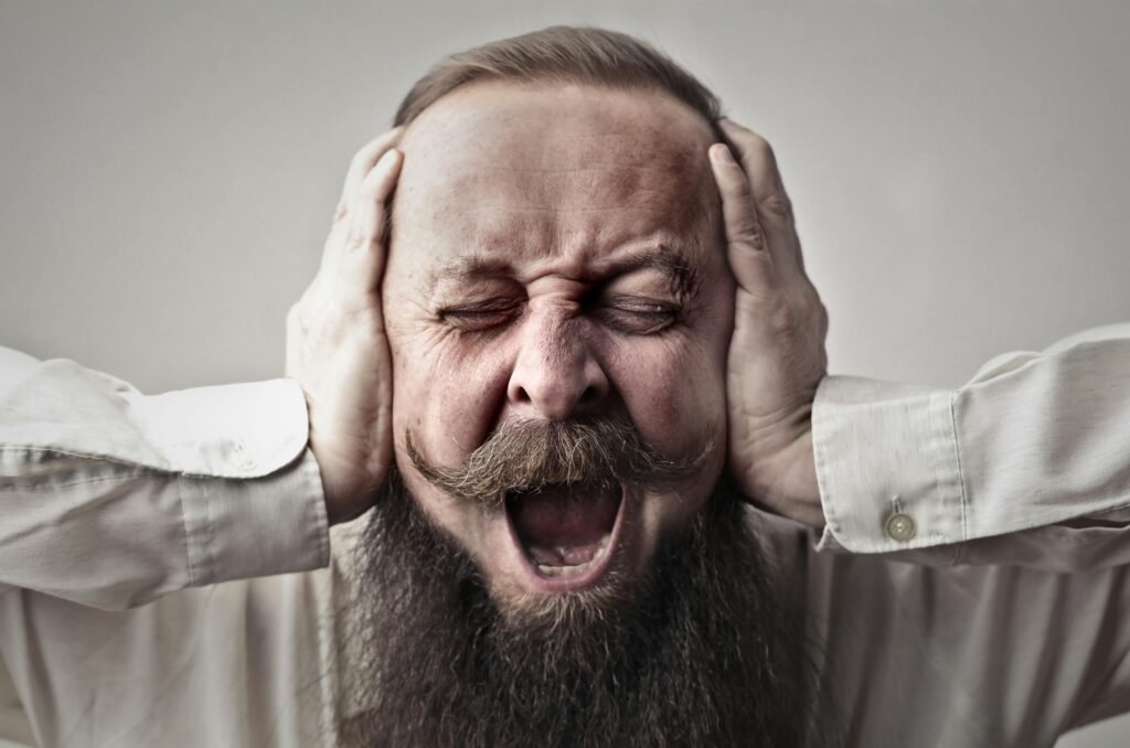 pexels-photo-3764535-3764535 Portrait of a man with facial hair, covering his ears and screaming, expressing stress or frustration.
