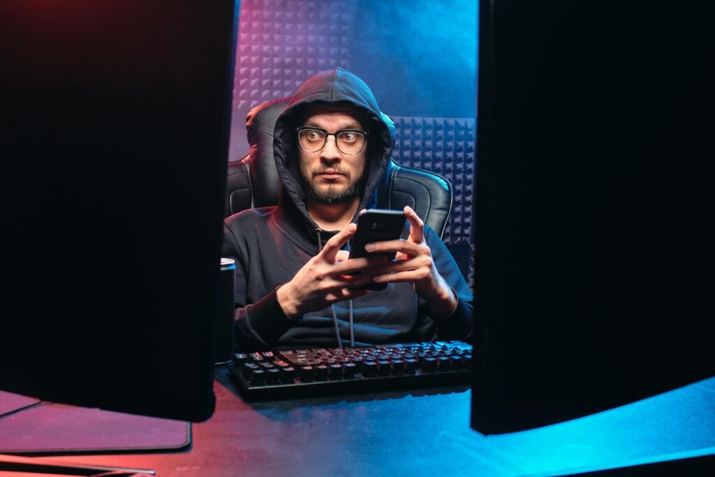 pexels-photo-6963062-6963062 A man in a hoodie using a smartphone, surrounded by tech gear in a dimly lit room.