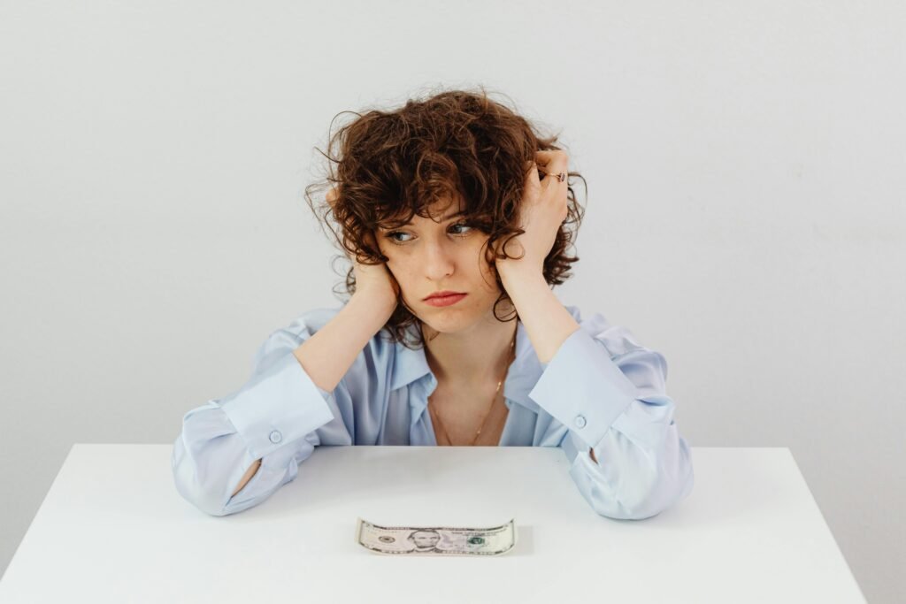 pexels-photo-7680366-7680366 A woman in a blue blouse looks worried with a dollar bill on a table, symbolizing financial stress.